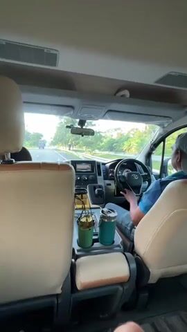 The video shows the interior of a van driving on a highway. The camera is positioned behind the driver and passenger seats, looking forward through the windshield. The driver, wearing a light blue shirt and a grey cap, is holding the steering wheel. Sunglasses are hanging from the center console, along with two travel mugs. The road ahead is visible, with trees and greenery on either side. Several trucks and cars are driving on the highway. The sky is bright, suggesting it is daytime. The van appears to be in motion, as evidenced by the blurred scenery and the perspective.