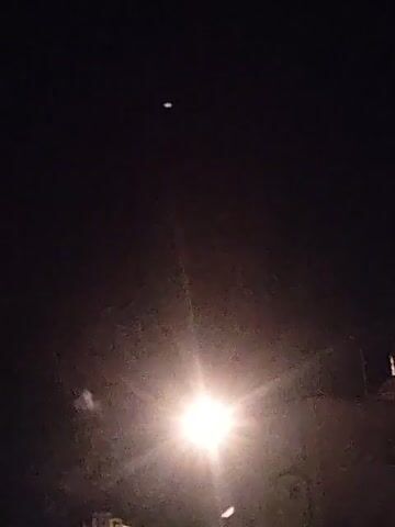 The video begins with a single, bright orange firework exploding in the dark sky, resembling a starburst. In the following frames, more fireworks appear, some in clusters and others individually, scattered across the black expanse. These fireworks are also orange and yellow, emitting sparks and trails of light. Below the fireworks, in the lower portion of the frame, the silhouette of buildings is visible against the night sky. The video captures the spectacle of a fireworks display.