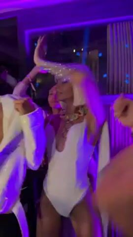 A video shows a woman with a blonde bob wig in a white swimsuit and a flowing white cape dancing in a club. She wears statement jewelry and has a jeweled arm cuff. The room has purple lighting, and other people are dancing nearby, but the focus is on her. One dancer wears a white shirt. Behind them is a paneled window. The music is upbeat.