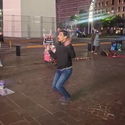 A man is dancing on a brick plaza at night. He is wearing a black jacket, blue jeans, and gray shoes, and has his hair in braids. He is standing in a wide stance and moving his arms and legs. To the left of him is a lit display and a person standing still. Behind him is a pink cart with various items on it and a man standing nearby looking at his phone. Further back is a street with cars driving on it, a pedestrian crosswalk, and an illuminated ferris wheel. Other people are standing and milling around in the background. The plaza is made of dark brick.