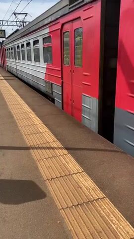 A train passes by a train platform. The train is red and grey. The platform has yellow tactile paving. The sky is overcast. The camera is static.