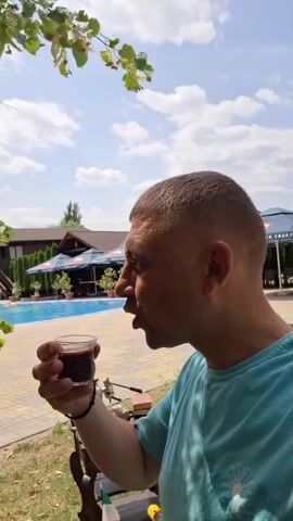 A man with a shaved head is sitting outside, taking a sip of a dark liquid from a small glass. He is wearing a turquoise t-shirt. In the background, there is a swimming pool and some buildings. The sky is blue with some clouds.