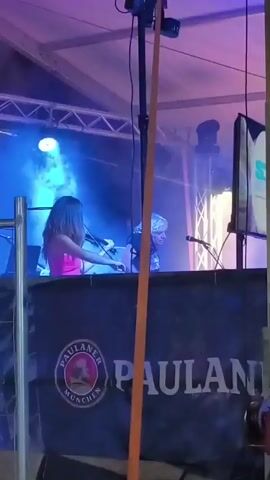 A woman in a pink top and a person with white hair in a black t-shirt are on a stage. The woman is holding a violin and bow, and the person with white hair appears to be operating DJ equipment. There are stage lights and smoke effects. A banner with the Paulaner logo is visible in front of the stage. A bright orange strap hangs down in the foreground, obscuring part of the scene.