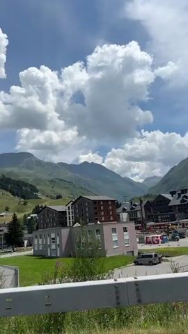 The video is a roadside view of a mountain village. In the foreground is a metal guardrail and some dry grass. The middle ground shows a modern building with a sloped, ribbed facade and a row of windows. Behind it are more buildings, some with multi-story structures and dark roofs, indicative of a resort or residential area. The background consists of lush green mountains under a bright blue sky with dramatic, puffy white clouds. The scene appears to be captured from a moving vehicle, as evidenced by the slight blur and shifting perspective.