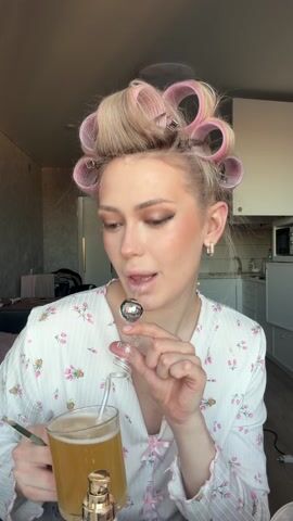 A fair-skinned woman with blonde hair in curlers is in a room that seems to be her apartment. She's wearing a light floral-patterned pajama top and square gold earrings. She holds a clear glass mug filled with a light amber liquid and a silver swizzle stick with a spherical end. She first stirs the liquid with the swizzle stick, then puts the spherical end of the swizzle stick in her mouth, looking directly at the camera before taking a sip from the mug. A kitchen area with a microwave and sink can be seen in the background.