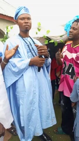 Here is a forensic description of the video:

The video shows a man in a light blue traditional Nigerian outfit speaking into a microphone. He's wearing a matching cap and a black beaded necklace. He stands surrounded by other men, some in traditional attire and others in casual clothing. The setting appears to be an outdoor event, possibly a wedding or celebration, indicated by the white drapery and decorative elements in the background. Some people are holding up the draped ceiling in the background. The man with the microphone is likely giving a speech or making an announcement. He seems to be speaking with enthusiasm.