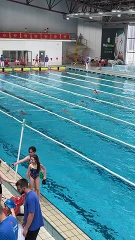 The video shows an indoor swimming pool during a swimming competition. Several children and adults are visible. A group of children in swimsuits stand near the edge of the pool, while others swim in the lanes. Several adults appear to be coaches or officials. The pool has multiple lanes marked by white lane dividers. Flags of different nations are displayed above the pool. A digital scoreboard displays names and times.  The environment appears well-lit and spacious.