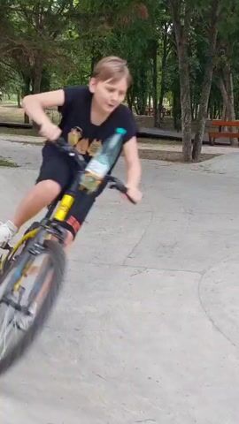In a skatepark setting, three young people are riding bicycles. The scene appears to be outdoors, with numerous trees and lush greenery forming the background. The weather seems fair, and the lighting suggests it's daytime, possibly late afternoon or early evening given the ambient light. The skatepark itself is constructed from smooth concrete, featuring ramps and curves, characteristic of a place designed for wheeled sports.

The central figure is a boy wearing a black t-shirt and black shorts, riding a bright yellow bicycle. He is captured in motion, his body angled as he navigates the skatepark terrain. To his right, another person on a bicycle is visible, though less prominently. Further in the background, a third individual on a bicycle is also seen. The overall mood of the clip seems to be one of activity and recreation, with the subjects engaged in enjoying their bicycles in a park environment. There is no visible text in the media. The location context of Lermontov, Russia, places this recreational activity within that specific geographic area.