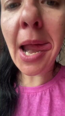 A woman with dark hair and green eyes is shown in a close-up shot, wearing a pink t-shirt. She is making a face with her tongue sticking out, then brings a fork with food to her mouth, her eyes wide. The background is blurry, but appears to be an indoor setting with a window to the left.