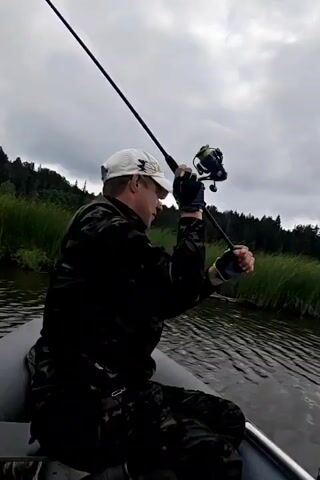 A man wearing a camouflage jacket and a white baseball cap sits in an inflatable boat and fishes. He holds a fishing rod and reel. The boat is on a body of water, and reeds and trees line the distant shore under a cloudy sky.