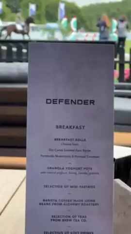 Here is a concise description of the video:

The video shows an outdoor seating area, likely at a restaurant or viewing lounge, with a view of a horse show jumping event. The focus is on the foreground, which features a table with a "Defender" breakfast menu and a bottle of Heinz ketchup. Behind the table, there are black outdoor chairs with gray cushions, and a partial view of an umbrella support. The background displays a horse show with a horse and rider jumping over an obstacle. There are a few spectators standing along the perimeter fence, observing the event. The day appears overcast.
