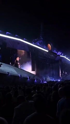 A person stands on a brightly lit stage, holding a guitar and singing into a microphone. The stage is elaborate, with large structures and lighting. A crowd is visible in the foreground, mostly in shadow. The overall scene is dark, suggesting it is nighttime and the event is an outdoor concert.