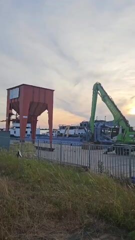 The video pans across a waterside industrial area at dusk. In the foreground, tall dry grass lines a metal fence. Beyond the fence, a large green excavator with an extended boom is positioned near a body of water. Several barges or boats are docked or moored in the water. To the left of the excavator is a large, red, industrial silo-like structure with a wide opening at the bottom. The sky above is a light blue with wispy clouds, and the sun is setting on the horizon, casting a warm glow. The overall impression is one of ongoing industrial activity in a port or harbor setting.