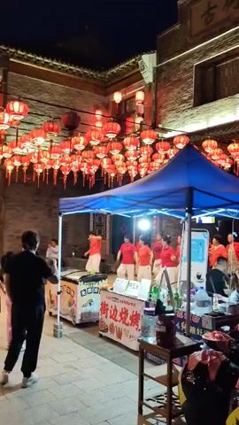 A group of people are dancing under a tent adorned with red lanterns at night. The dancers are wearing red tops and white bottoms. The tent is set up in an outdoor market area with stalls selling food. Spectators are watching the performance. There are also decorative lights and architectural elements that suggest a traditional setting.