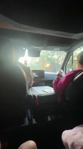 The video, shot from the back seat of a moving vehicle, features two people in the front seats, one driving and the other seemingly using a phone. The driver, clad in a pink sweater, is visible from the shoulders up, while the person in the passenger seat is only partially visible, wearing a light-colored shirt. The dashboard of the vehicle has a small screen and some money and coins are on the console between the seats. Outside, the sun is low in the sky, creating a bright glare through the windshield as they drive past trees. A speed limit sign is visible, indicating a speed of 70.