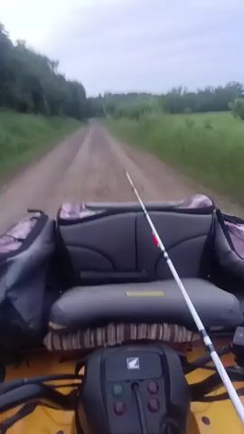 The video shows a first-person view from a yellow Honda ATV traveling on a dirt road. A fishing rod is mounted in front of the back seat. The road is bordered by green grass and trees on both sides. The sky is overcast.