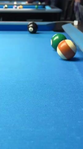 The video shows a billiard table with a blue felt surface. Several billiard balls are visible, including a black 8-ball positioned near the back of the table, and a striped orange and white ball, a solid green ball, and a white cue ball in the foreground. The cue ball is in motion, blurring as it moves. A metallic rail runs along the side of the table. In the background, other billiard tables are visible, along with people standing and observing. The camera is positioned at table level and remains static throughout the video.
