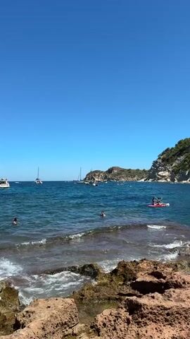 The video shows a clear blue sky and a bright sunny day at a coastal location. The water is a vibrant blue, with small waves breaking on a rocky shoreline in the foreground. In the middle ground, several people are swimming and enjoying the water. Further out, there are sailboats and a small motorboat. On the right side of the frame, a person is paddling a red kayak or paddleboard. The background features a lush green hillside with some buildings visible at the top. The overall scene depicts a tranquil and enjoyable day at the beach.