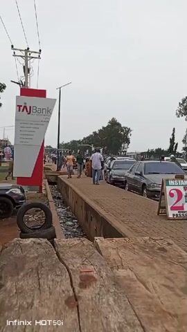 The video shows a street scene in what appears to be a developing country. There is a sidewalk beside a road with several cars parked along it. People are walking along the sidewalk. A sign advertises "Instant 2 Minutes Passport" photo services. There is a TAJ Bank sign in the background. Debris is visible in a narrow trench or drainage ditch beside the sidewalk. Old tires are stacked near the foreground. The sky is overcast.