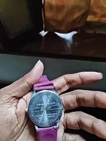 A person's hand holds a wristwatch with a purple band. The watch face is dark blue with white numbers and hands. There is a small, light-colored object on the watch face, possibly dirt or debris, near the center. The background is dark and out of focus, with hints of a patterned fabric. The watch is being rotated, showing both the back of the watch case and the front face.