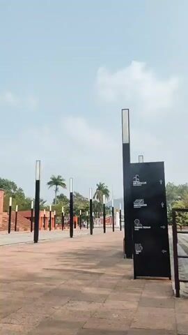 A paved outdoor area with a clear sky above. In the foreground, a tall black post with a white light fixture stands to the right. Adjacent to it is a black directional sign with white icons and text. The pavement is made of rectangular tiles in shades of orange and brown. A row of similar black posts with white light fixtures stretches into the distance. In the background, there are buildings constructed of red brick with stepped seating areas. Green trees and palm trees are visible behind the buildings, and a hill or mountain range can be seen in the far distance. The overall impression is of a public space or monument.