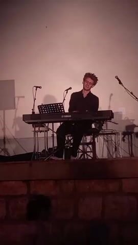 A young man with light brown, tousled hair is playing a black Yamaha keyboard. He is wearing a dark collared shirt and black pants. The keyboard is on a black metal stand, with a music stand in front of it. Two microphones are positioned on stands behind the keyboard. The background is a plain, light-colored wall, possibly a projection screen. The man is seated on a stool. Below the stage is a textured wall made of large, rough stones. In the foreground, out of focus, is the silhouette of a person's head. The lighting is dim, with a pinkish-orange hue. The video appears to be taken from a slightly low angle, looking up at the stage.