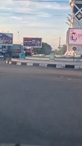 A daytime outdoor scene shows a paved road with a roundabout in the background. Two cyclists are riding on the road in the foreground, with one on the left and another on the right. The cyclist on the left is wearing a helmet. The cyclist on the right is wearing dark clothing and is partially obscured. Billboards are visible behind the roundabout. One billboard on the left shows people in yellow shirts. Another billboard in the center has text in red and black, and displays images of cigarette packs. A large digital billboard on the right shows a pink advertisement with a person's face. The sky is partly cloudy with the sun setting. The environment appears to be a town or city intersection.