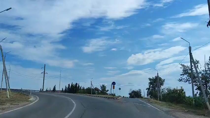 The video shows a view from inside a vehicle driving on a paved road under a cloudy blue sky. The road curves to the left. On the right side of the road, there are utility poles with wires, streetlights, trees, and a blue circular traffic sign with white arrows indicating a curve to the right. A yellow and black striped sign is also visible. The landscape appears to be rural or semi-rural, with some buildings visible in the distance. The video captures the movement of the vehicle along the road.