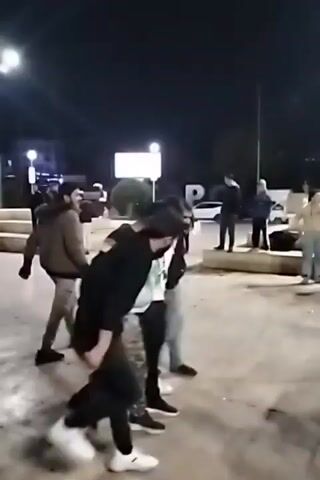 The video shows a group of four men in an urban outdoor setting at night. They are engaged in a synchronized dance, moving in a line with coordinated steps and gestures. Other people are standing around watching. The area is lit by streetlights and buildings in the background have some lights on as well. The ground appears to be a paved surface. The men are dressed in casual clothing.