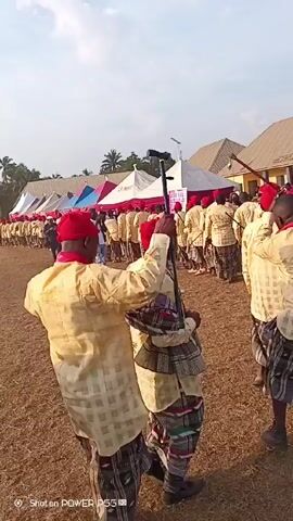 A large group of people, mostly men, is walking in a line across a grassy field. The people are wearing matching outfits consisting of red caps, beige patterned shirts, and patterned wraps around their waists. Many of them are carrying black canes or staffs, some of which are decorated with red fabric. In the background, there are several white tents and some low buildings with peaked roofs. The sky is bright and clear. A watermark on the bottom left corner reads "Shot on POWER P55." The group appears to be participating in a procession or cultural event.