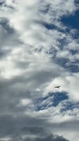A small, single-engine airplane is flying in the sky. The sky is partly cloudy, with patches of blue visible between the clouds. The plane appears to be a dark silhouette against the sky.