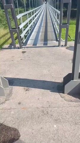 A first-person perspective video shows a person riding a bicycle across a suspension bridge. The bridge has a metal walkway with railing on both sides. The surrounding environment is green and appears to be a park or natural area. Sunlight casts shadows on the bridge as the cyclist moves forward.