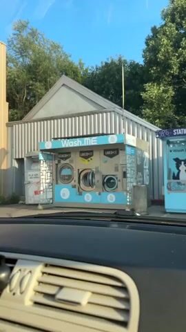 A self-service laundromat is seen from the perspective of a car. Three washing machines are visible, one labeled "SÉCHE 20KG" (dryer), and two labeled "LAVE 20KG" (washer). The middle washer has its door open. The laundromat is a metal structure with a pitched roof and a blue and white awning that reads "Wash.ME". Trees and a blue sky are visible in the background.