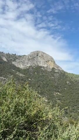 The video is a landscape shot of a mountainous region with lush green vegetation covering the slopes. In the foreground, there is a dry, grassy area with some bushes and a stone wall bordering a paved road. The road curves to the right as it moves away from the viewer. In the background, a large, imposing gray rock formation dominates the scene, with a rugged, cliff-like face. Further in the distance, more mountain ranges can be seen, fading into the hazy horizon under a bright blue sky dotted with white, wispy clouds. The sun appears to be shining, casting a natural light on the landscape. The camera pans slightly to the right, revealing more of the winding road and the expansive mountain vista.