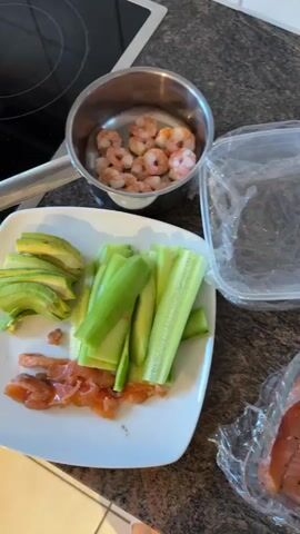 The video displays a countertop with various ingredients for preparing food, likely sushi or a similar dish. The items visible include a pot of cooked rice, peeled shrimp in a small metal bowl, sliced avocado, julienned cucumber, and thinly sliced smoked salmon. There are also two small containers of cream cheese and a piece of salmon skin on a paper towel. A clear plastic container with plastic wrap is also present, suggesting ingredients are being organized or prepared for assembly. The background shows a kitchen counter and what appears to be the edge of a stovetop. The lighting is bright, indicating an indoor setting, likely a kitchen. The video appears to be a close-up, top-down view of the food preparation area.