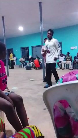 The video shows a room with light blue walls and a light-colored concrete floor. Several people are in the room, sitting on white plastic chairs and standing around. A young man in a white long-sleeved shirt with a graphic on it and black pants is standing in the center of the room. Another young man wearing a pink short-sleeved shirt stands next to him, they appear to be talking. Several metal support posts are visible throughout the room. There are windows on the far wall.  Various items are scattered on the floor. The camera is held by someone sitting in a chair, offering a partial view of their legs and the chair. The scene seems to be indoors, possibly a social gathering or event.