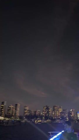 The video shows a blurred cityscape at dusk, likely taken from a moving boat. The sky has a mix of dark blues and pinkish hues. Silhouetted figures are visible in the foreground, suggesting people on the boat. Music is playing in the background.