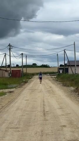 A person walks down a dirt road. Another person, holding a soccer ball, walks towards the first person, extending their arm in a waving motion. The road is lined with utility poles and power lines. On the left side of the road are fences and a building. On the right side of the road are more buildings and a speed limit sign. The sky is overcast with dark clouds. The ground is covered in dirt and some grass.