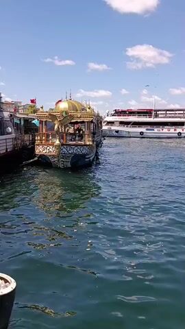 The video captures a sunny day with a blue sky and scattered clouds. The scene is set on the water, likely a harbor or bay, with calm, blue-green water filling the foreground and midground. In the left midground, a uniquely decorated, ornate boat with a gold dome and intricate patterns is docked. To its right, a larger, white passenger ferry with multiple decks and the name "TURYOL" visible on its side is also present. The ferry appears to be in operation, with its lights on and suggesting movement. In the background, a city skyline with buildings and what appears to be a bridge can be seen on the horizon. The lighting suggests it is daytime.