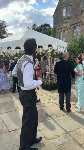 The video shows a man playing an accordion while walking across a stone patio. He is dressed in a white shirt, a grey vest, and pinstripe pants. He smiles and looks towards the camera as he plays. In the background, other people are mingling under a large white tent. A stone building with windows is visible behind the tent. There are also some bushes and pink flowers in the foreground and background.