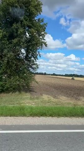 A tractor plows a field under a cloudy blue sky. The field is brown and tilled, with dry grass on the edges. There are trees and houses in the distance. The video is a landscape shot with the camera panning across the field.