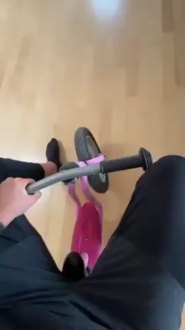 A person wearing black pants and socks is sitting on a small pink balance bike on a light-colored wooden floor. They are holding the handlebars and using their feet to push themselves forward. The camera is positioned above the person, looking down at the bike and their legs. The background includes glimpses of furniture and a doorway.