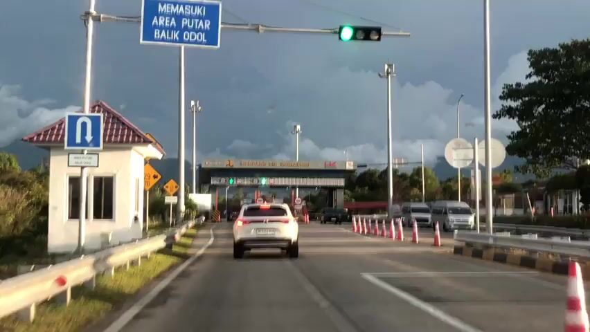 A white SUV approaches a toll plaza on a multi-lane highway. The SUV is in the rightmost lane, which is marked with orange cones. Other vehicles, including a black sedan and several vans, are in the lanes to the right of the SUV. The toll plaza is a large structure with multiple booths, some of which have green lights indicating they are open. The sky is cloudy and appears to be threatening rain. There are mountains in the distance. The SUV has a license plate that reads BK 1612 MEB. The video is shot from the perspective of someone inside another vehicle following the SUV.