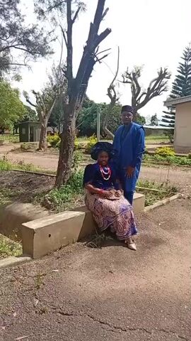 A woman sits on a concrete bench in front of a man who stands behind her. Both are dressed in traditional blue and patterned garments, and they are outdoors, with trees and buildings in the background. The woman wears a blue headwrap and a necklace, and the man has a beard and is wearing a hat. They appear to be posing for a photograph.