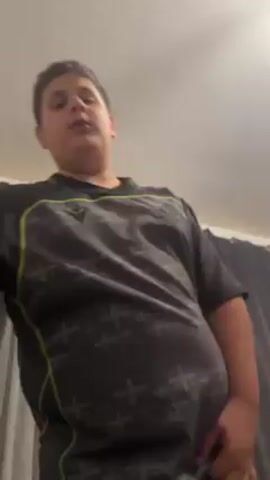 A young person stands indoors, facing the camera, wearing a patterned black and gray t-shirt. They put their hands behind their head and speak. Behind them are gray curtains and a window. The room is lit by an overhead light.