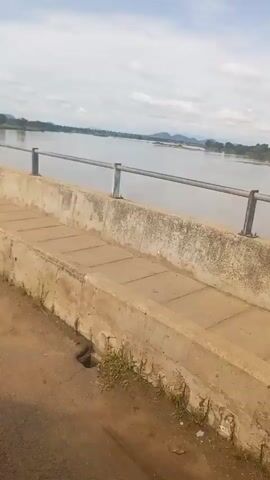 The video shows a view from a bridge overlooking a wide river. On the bridge's concrete barrier, graffiti is visible. The first portion shows the barrier with no visible graffiti. Then, "MCN PHILIP JOHN'S" is seen spray-painted on the barrier. After that, "BALT" is spray-painted on the barrier. Finally, a person in a light blue garment runs across the bridge, passing the graffiti. The river is calm and wide, with land and trees visible on the opposite bank. The sky is cloudy.