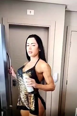 A woman with long dark hair and a tiger-print outfit is standing in what appears to be a kitchen. She is holding a white bottle and seems to be opening it. The room is well-lit, suggesting it's daytime. There are two doors visible in the background, one with a white handle and the other a plain grey door. A refrigerator is partially visible on the left side of the frame. The woman's expression is neutral, and her body language is relaxed. The overall scene is domestic and casual. There is no visible text in the media file.