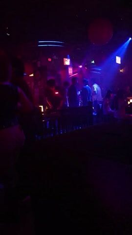 The video captures a dark, dimly lit nightclub environment with vibrant blue and pink neon lights. In the foreground, silhouetted figures are visible, suggesting a crowd of people. The central focus shifts to a stage or elevated platform where one or two individuals are performing or dancing. One performer, dressed in a light-colored top and dark shorts, is seen in a dynamic pose. Another individual in a dark outfit is positioned below. The lighting creates a pulsating and energetic atmosphere characteristic of a nightclub scene.