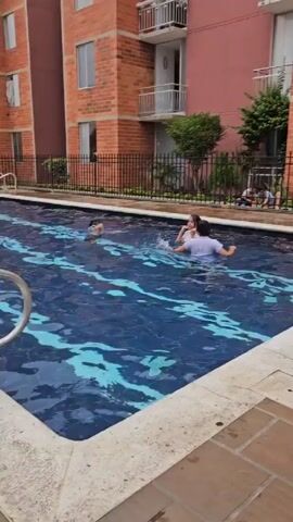 The camera pans across an outdoor swimming pool where several people are swimming. The pool is lined with dark blue tiles that give the illusion of water flowing underneath. The pool is surrounded by a stone-tiled deck. Beyond the pool, there is a black metal fence that separates the pool area from other outdoor spaces. Beyond the fence is a playground and a basketball court where other children are playing. Behind the playground and pool area are rows of tall, multi-story apartment buildings constructed of red brick. The sky above is overcast and gray.