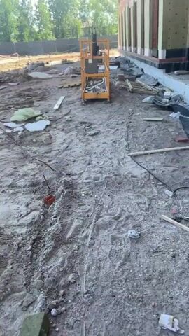 The video shows a construction site on a cloudy day. The camera pans across a muddy area with construction debris, including wood, pipes, and insulation. A yellow lift is visible in the distance. The camera then moves to a building under construction, where workers are installing insulation on the exterior walls. The insulation is light brown and has red circular markers. The building is made of brick and concrete. Another section of the building shows exposed red brick walls with metal reinforcements. More construction equipment, including a black control box and blue buckets, is visible near the building. Two workers are seen wearing hard hats and safety vests, one in an orange shirt and the other in a blue shirt, actively working on the building's facade. The ground in front of the building is a mixture of dirt, gravel, and construction materials.